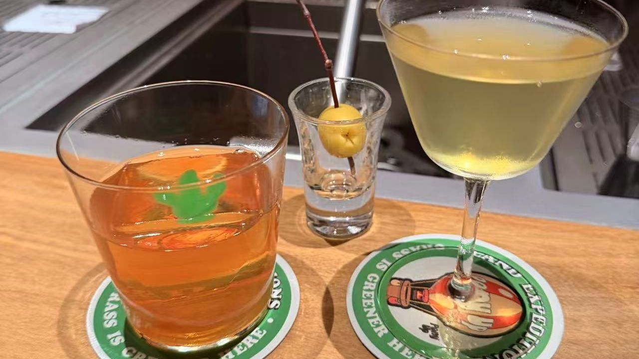 An old fashioned and a martini on green paper coasters at a wooden bar counter.